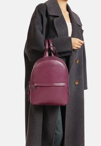 Zaino in pelle borgogna con forma arrotondata, maniglie doppie e tasca frontale. Indossato sopra un cappotto grigio, che mette in evidenza il suo design elegante.