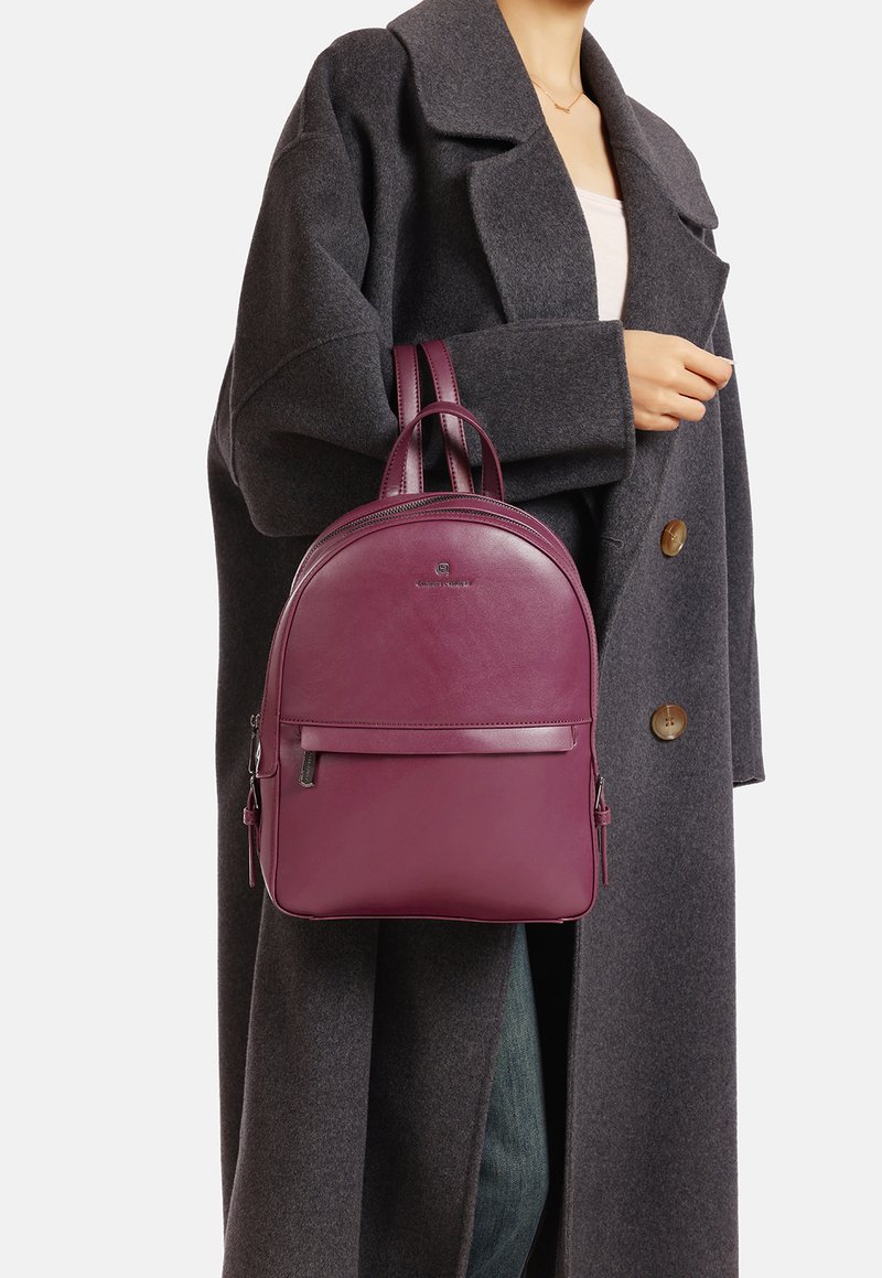 Zaino in pelle borgogna con forma arrotondata, maniglie doppie e tasca frontale. Indossato sopra un cappotto grigio, che mette in evidenza il suo design elegante.