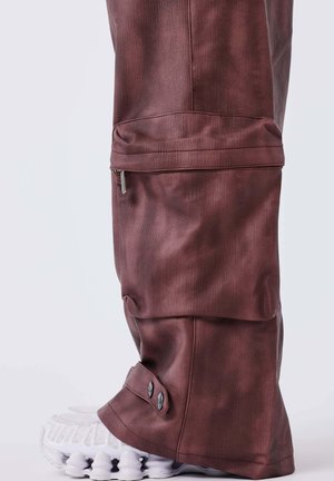 Pantalons en tissu texturé bordeaux foncé, avec une poche zippée et des accents de bouton au niveau de l'ourlet, associés à des baskets blanches.