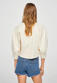 Chaqueta de crema recortada con mangas anchas, hombros estructurados y tela texturizada, combinada con shorts de denim azul que tienen bolsillos traseros.