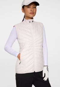 Lätt, ljusrosa zip-up väst med ett quiltat mönster, hög krage och två sidofickor, bärs över en långärmad vit topp.