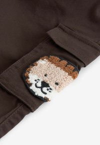 Brauner Stoff mit einem plüschigen Löwenpatch; das Patch zeigt Farben in Braun, Creme und Schwarz; Details umfassen strukturierte Fasern und gestickte Merkmale.