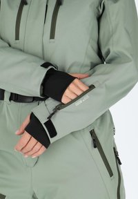 Light green insulated outerwear with black accents, featuring a high collar, adjustable cuffs, and multiple zippered pockets.