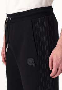 Black jogger pants with a textured side panel, drawstring waist, and a small logo patch on the thigh. Fabric appears soft and elastic.