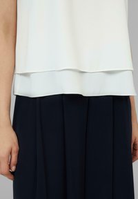 Witte gelaagde blouse met een gladde stof, luchtige drape en een marineblauwe geplooide rok. Zichtbare hand nabij de zoom van het kledingstuk.