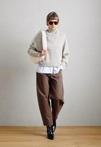 Beige pletený rolák, přes světle modrou košili, s hnědými širokými kalhotami a černými kotníkovými botami, nese bílou plyšovou tašku.