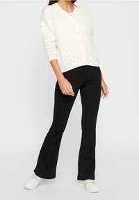 Kvinna i krämfärgad knapprad cardigan, svarta utsvängda byxor och vita sneakers som står mot en enkel bakgrund.