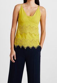Camisole jaune avec un décolleté en V, dotée d'une finition en dentelle festonnée au bas. Les bretelles sont fines et le tissu a une texture lisse.
