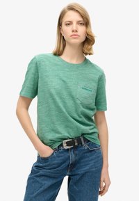 Grønt meleret bomulds T-shirt, rund halsudskæring, korte ærmer, brystlomme med "SUPERDRY" tekst, parret med blå denim jeans og et sort bælte.