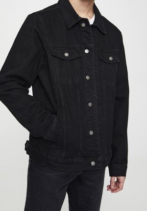 Osoba ubrana w czarną kurtkę dżinsową z srebrnymi guzikami i kieszeniami z przodu, w połączeniu z czarnymi dżinsami i białą koszulą pod spodem.