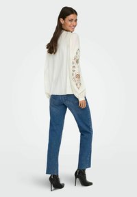 Biała bluzka z koronkowymi detalami na rękawach, zestawiona z niebieskimi spodniami typu high-waisted i czarnymi botkami na wysokim obcasie, widok od tyłu.