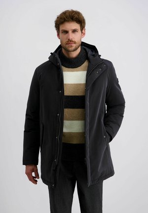 Schwarze, wasserfeste Parka mit Kapuze, ausgestattet mit einem Reißverschluss und Druckknöpfen, getragen über einem gestreiften, gestrickten Pullover in neutralen Farbtönen.