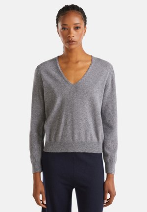 Vrouw in een grijze V-hals lange mouw trui en donkerblauwe broek, staand met ontspannen armen tegen een eenvoudige witte achtergrond.