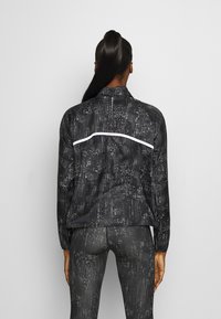 Chaqueta atlética negra con un patrón geométrico reflectante, que cuenta con un alto cuello y una franja de acento blanca en la parte posterior.