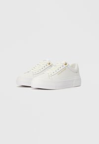 Lage witte sneakers met dikke zolen, witte veters, gouden oogjes en "Tommy Hilfiger" branding aan de zijkant.