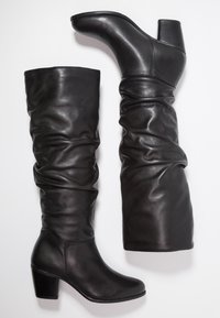 Bottes en cuir noir montantes avec un design décontracté, un talon carré et une texture lisse. La tige a une coupe relâchée, présentant des plis subtils.