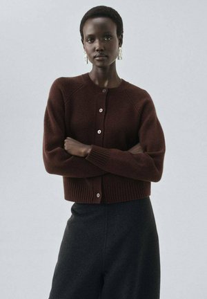 Gilet en tricot marron avec un col rond, un ourlet côtelé et des boutons. Le modèle le porte avec un pantalon noir taille haute.