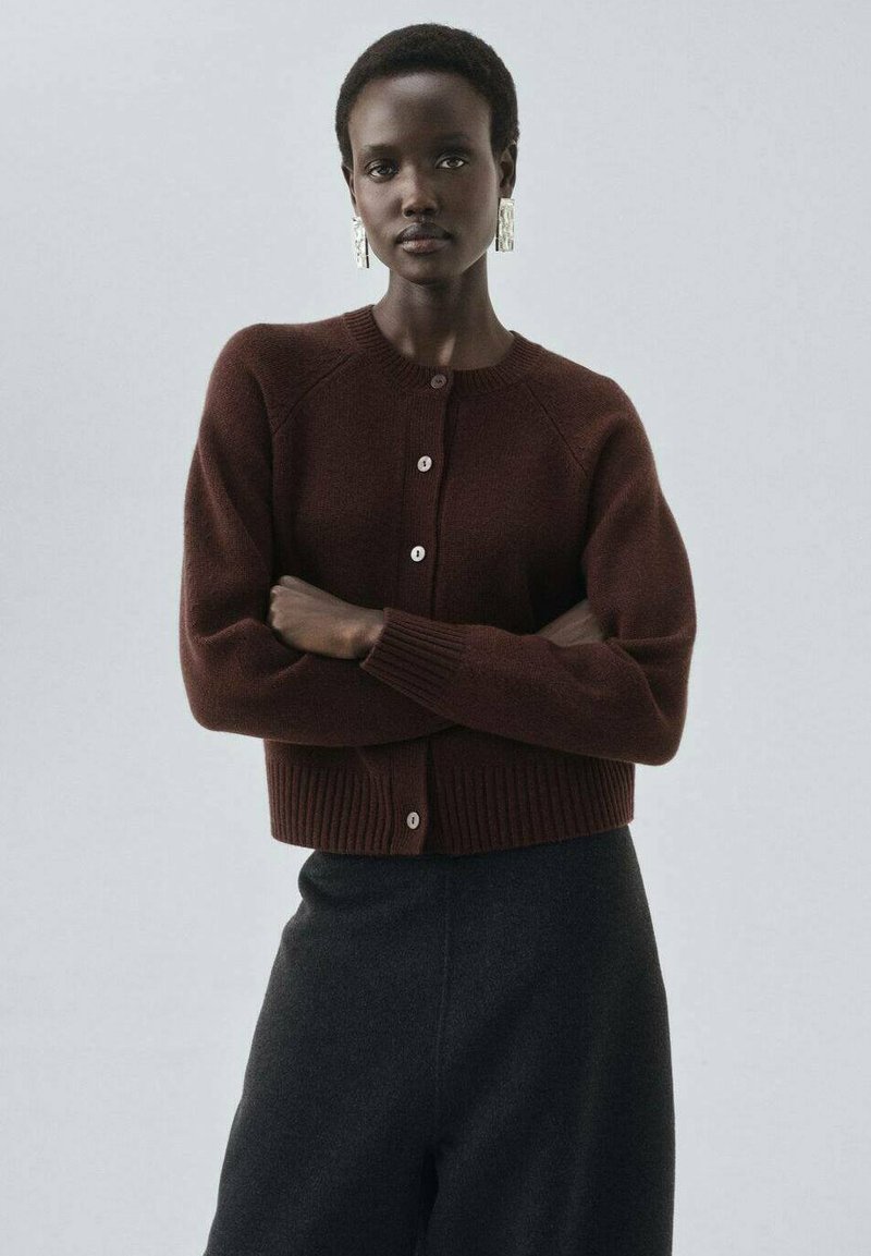 Gilet en tricot marron avec un col rond, un ourlet côtelé et des boutons. Le modèle le porte avec un pantalon noir taille haute.