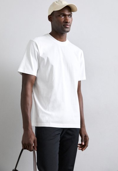 Camiseta de algodón blanca con mangas cortas y cuello redondo; modelo sosteniendo un bolso marrón, llevando una gorra beige clara, de pie contra un fondo gris.