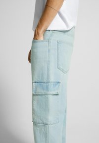 Světle modré cargo kalhoty vyrobené z denimu, se boční kapsou, zadními kapsami a volným střihem. Tkanina má mírnou strukturu.