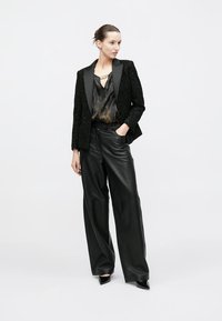 Zwarte textuurblazer met satijnen revers, gecombineerd met een donkerblouse met patroon en wijde zwarte leren broek. Zwarte schoenen met een puntige neus.