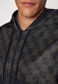 Chaqueta negra con capucha y un patrón a cuadros en verde oscuro y dorado. Presenta un cierre con cremallera y un detalle de cordón en la capucha.