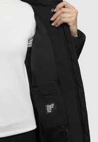 Giubbotto nero trapuntato con fodera interna liscia, dotato di tasca a toppa e etichetta con logo. Il modello indossa un maglione bianco sotto.