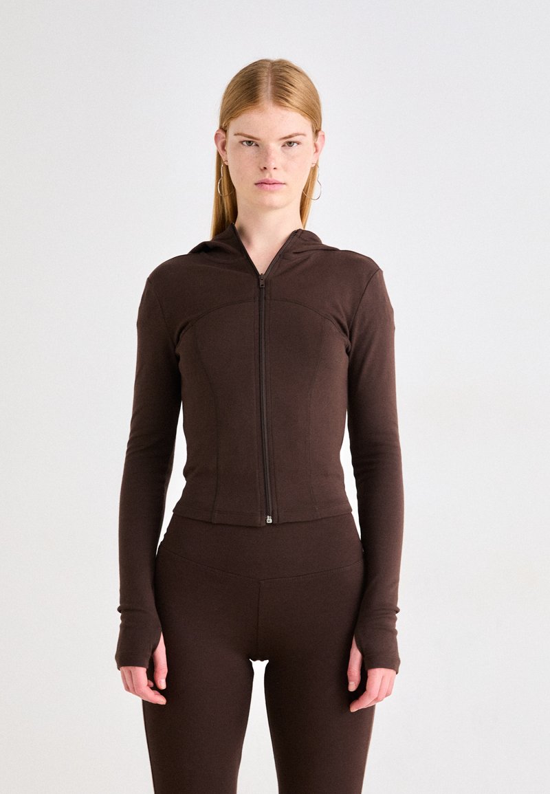 Sudadera con cremallera marrón en tejido elástico suave, diseño ajustado, mangas largas con agujeros para los pulgares y capucha. Combinada con leggins marrones a juego.