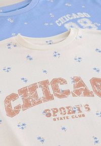 Dwie koszulki, niebieska i kremowa. Niebieska koszulka ma kwiatowy wzór oraz napis "CHICAGO 88". Kremowa koszulka posiada kwiatowy wzór i nadruk "CHICAGO SPORTS STATE CLUB".