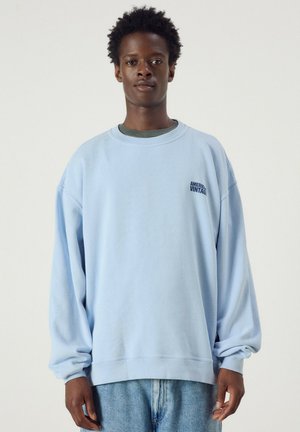 Giovane uomo con capelli corti e ricci che indossa una felpa oversize azzurra e jeans blu, in piedi davanti a uno sfondo semplice.