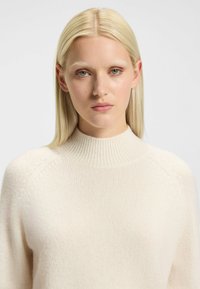 Pullover crema chiaro realizzato in morbido tessuto a maglia. Collo alto a coste, cuciture con spalle a caduta e vestibilità rilassata. Nessun motivo visibile.