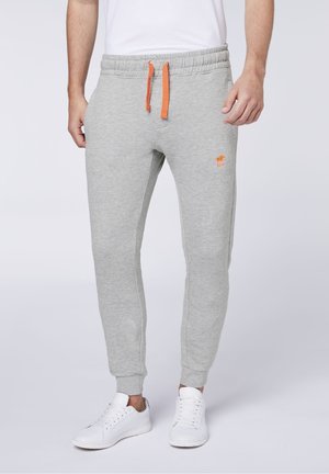 Ein Mann trägt hellgraue Jogginghosen mit orangefarbenen Kordeln und Logo, kombiniert mit weißen Sneakers, und steht vor einem schlichten Hintergrund.