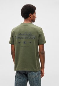 Oliivinvihreä puuvillainen lyhythihainen t-paita, jossa on laivastonsininen painatus, jossa lukee "stockholm ATHLETES CLUB 2015" ja ympyrälogo takana.