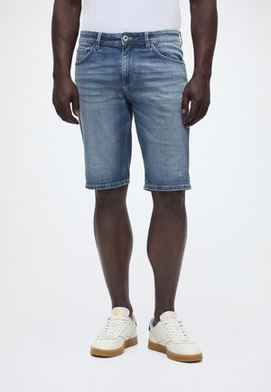 Mężczyzna w niebieskich szortach z denimu o długości do połowy uda, w białych tenisówkach z gumową podeszwą, stojący na tle jednolitej białej ściany.