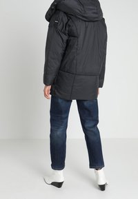 Černá prošívaná bunda s kapucí, hladkou texturou a viditelnými švy, doplněná tmavými džínami a bílými kotníkovými botami.