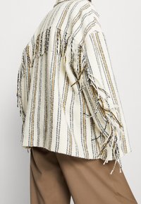 Giacca frangiata a righe realizzata in tessuto intrecciato con tonalità di crema, grigio e giallo. Presenta una vestibilità morbida e bordi grezzi e texture.