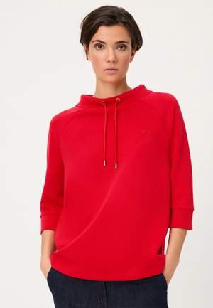 Woman with short dark hair wearing a bright red pullover with drawstrings and dark jeans, standing with hands in pockets.