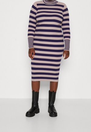 Frau trägt ein midi-langes, langärmliges Kleid mit lila und beige horizontalen Streifen und schwarzen, klobigen Stiefeletten vor einfarbigem Hintergrund.