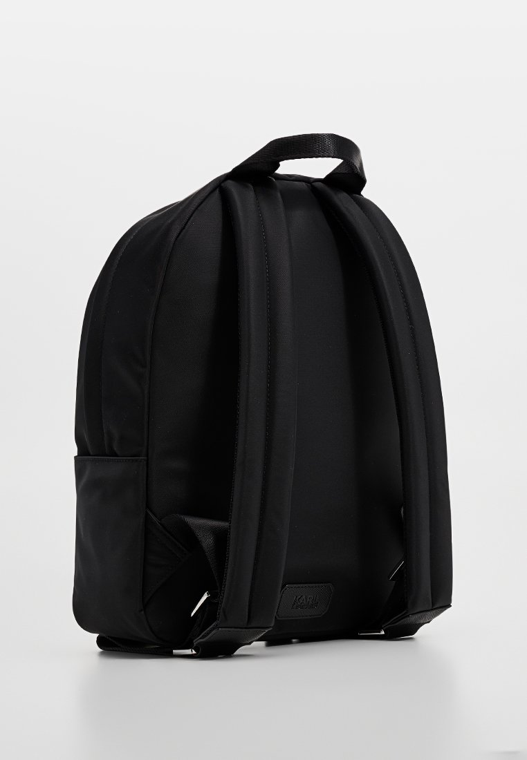 Sac à dos en tissu noir avec des bretelles rembourrées, une poche latérale et une petite étiquette de marque. Texture lisse et design minimaliste.