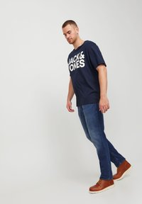 Άνδρας φορώντας μπλούζα navy blue Jack & Jones, μπλε τζιν και καφέ δερμάτινες μπότες, ποζάροντας μπροστά σε απλό ανοιχτό φόντο.