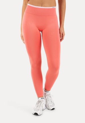 Mujer con leggings coral de cintura alta y zapatillas blancas con calcetines tobilleros estampados con logo, de pie contra un fondo claro y liso.