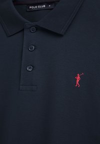 Polo blu navy con tre bottoni, piccolo logo ricamato di un giocatore di polo rosso sul petto e etichetta "POLO CLUB" all'interno del colletto.