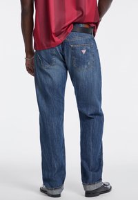 Vaqueros de mezclilla azul con un ajuste holgado, costuras visibles y una sutil textura desgastada. Presenta un logo en forma de triángulo en el bolsillo trasero.