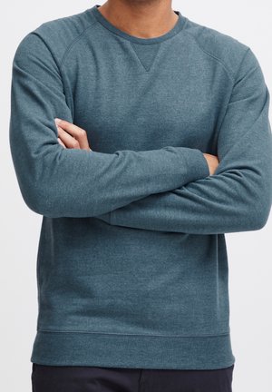 Mand i teal crewneck sweater med armene krydset, vist fra skuldre til hofter mod ensfarvet baggrund.