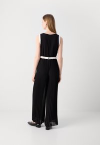 Combinaison noire plissée avec un détail de ceinture blanche, sans manches, en tissu lisse et avec une fermeture à l'arrière. Des ballerines complètent la tenue.