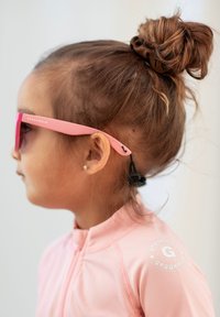 Vaaleanpunaiset aurinkolasit, joissa on geometrinen muotoilu, joita kantaa lapsi. Asusteena on vaaleanpunainen tekstuuripintainen takki, jossa on brändäys olkapäällä.