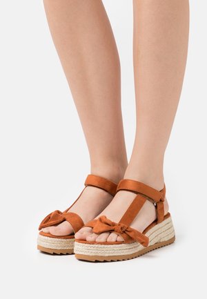 Brown wedge sandals with ankle straps and front bow detail worn on bare feet, showing textured rope platform soles.