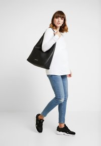 Bolsa tote de couro preta com uma forma arredondada, apresentando uma textura suave e branding discreto, combinada com uma blusa de manga longa branca e jeans azuis.