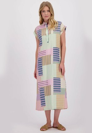 Vrouw staat in een pastelgekleurde patchwork midi-jurk met korte mouwen en slip-on tan sandaaltjes voor een effen witte achtergrond.