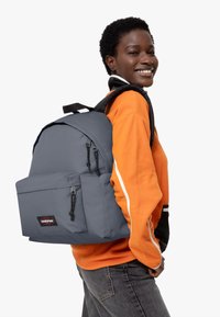 Sac à dos gris avec deux poches zippées, bretelles ajustables et logo Eastpak. Porté sur un vêtement noir et orange. Le tissu semble durable.